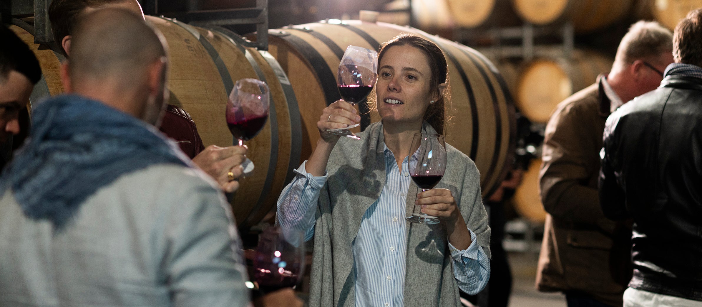 Woman holding a glass of red wine in a winery setting with other people and barrels.
